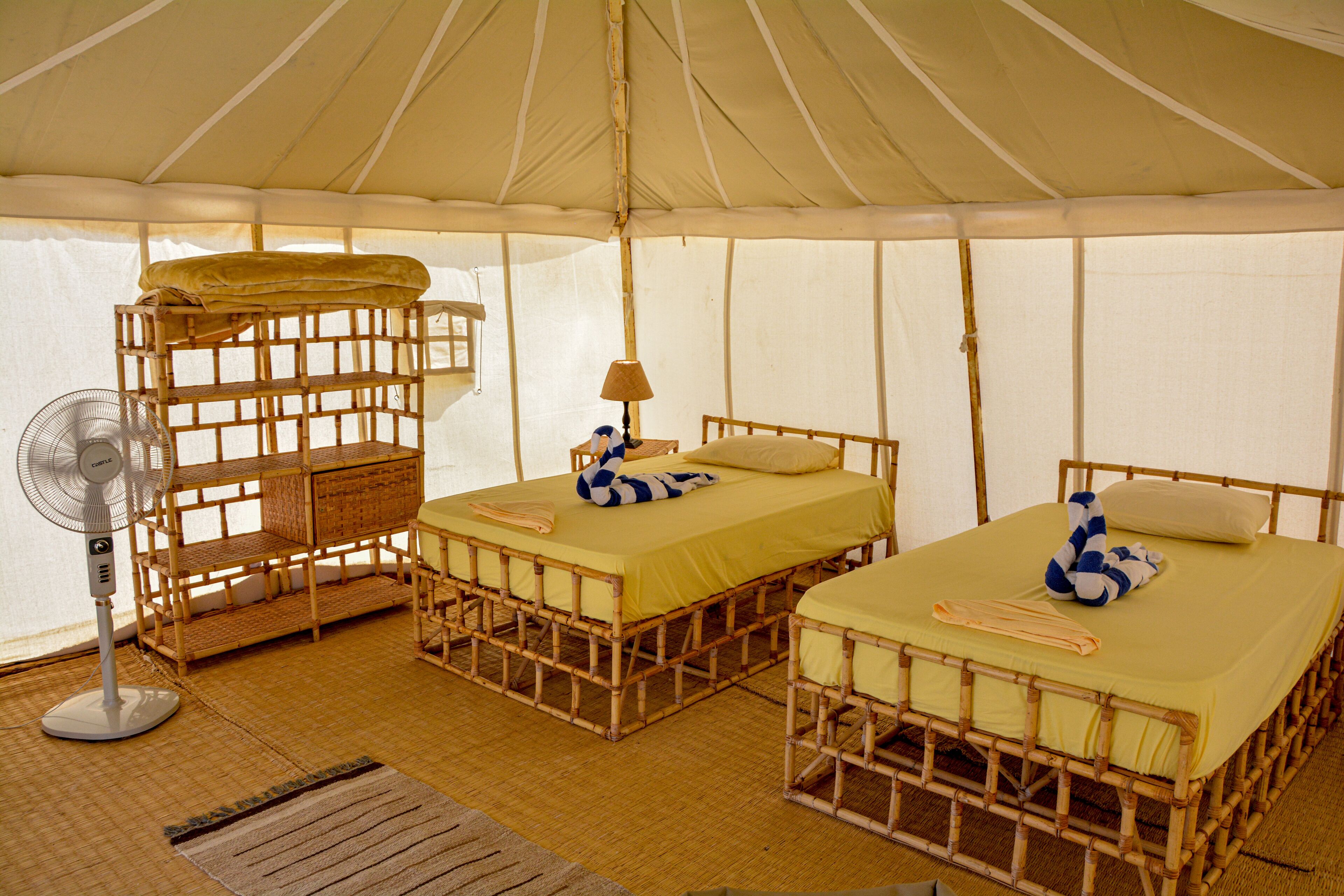 A tented accommodation with two beds, a fan, and a woven bench.