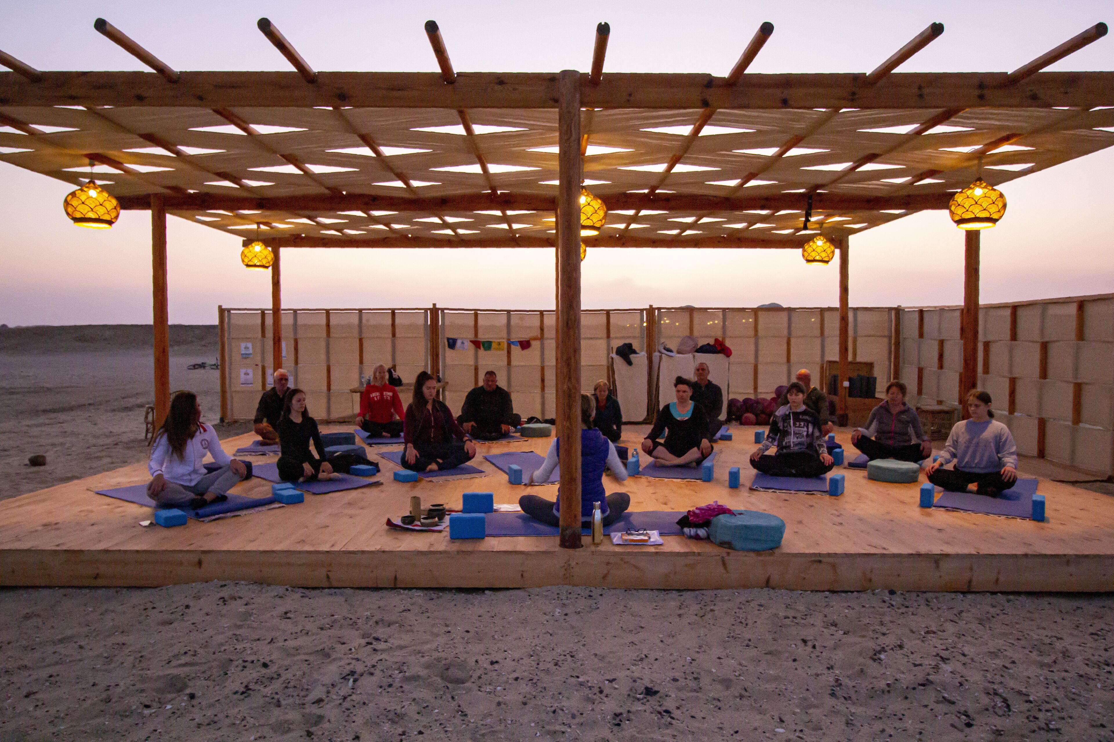 A group of people sitting on mats under a wooden pergola with hanging lights, on a sandy beach.