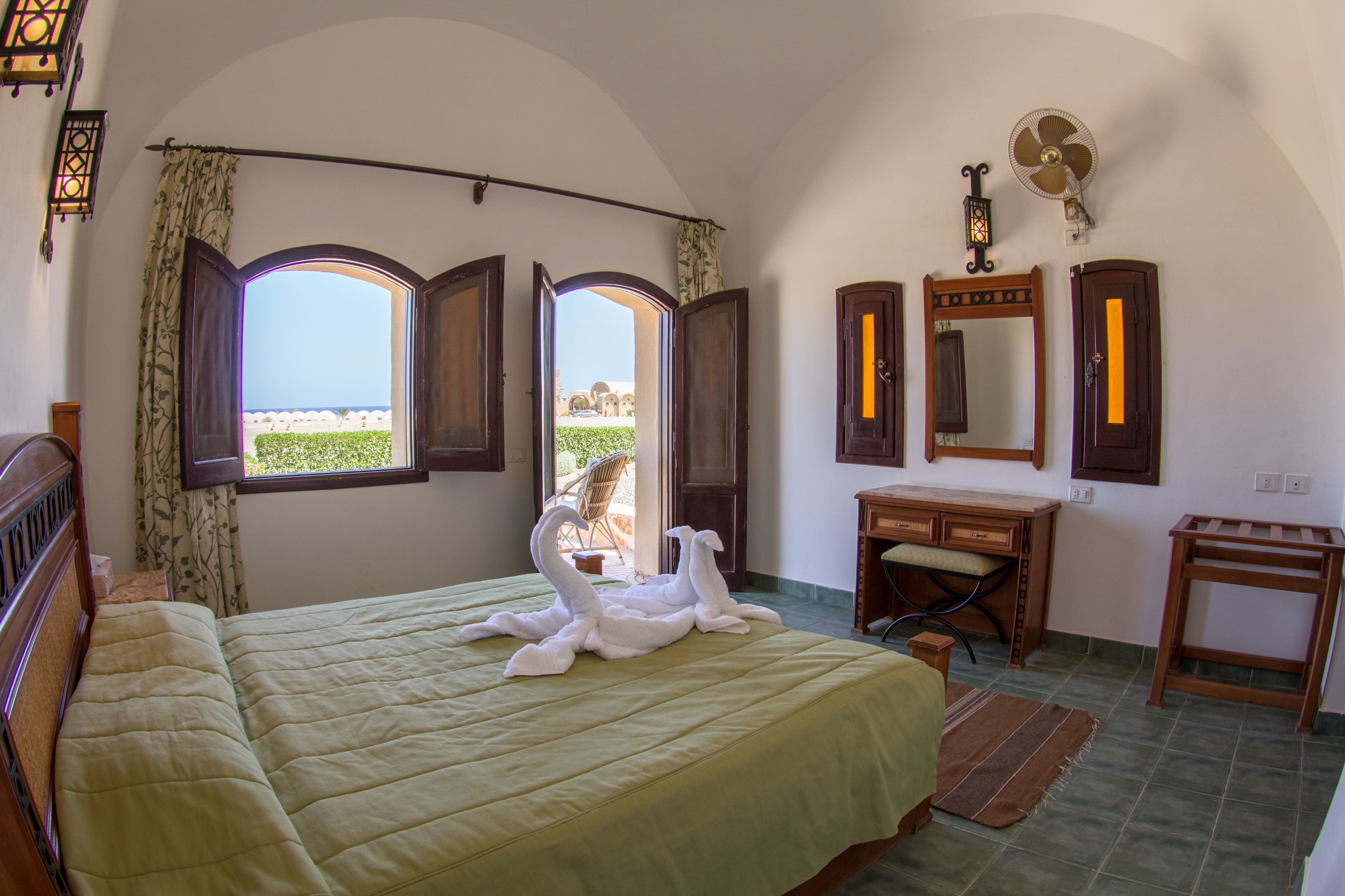 A bedroom with a bed, two open windows, a dresser, a vanity, and a fan.