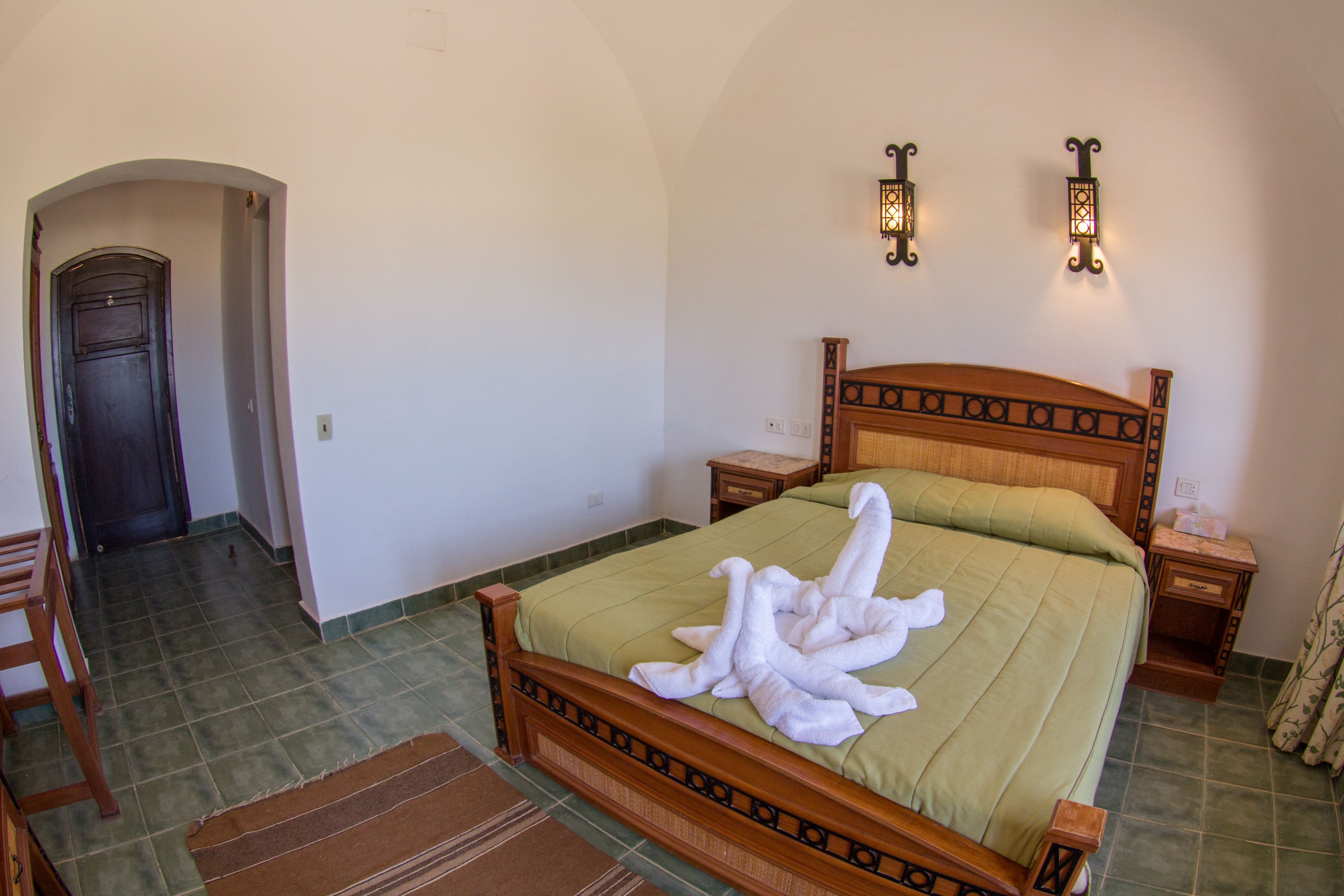 A neatly made bed with a green bedspread and a white towel arrangement. There are two bedside tables with lamps, a wooden headboard, and a door in the background.