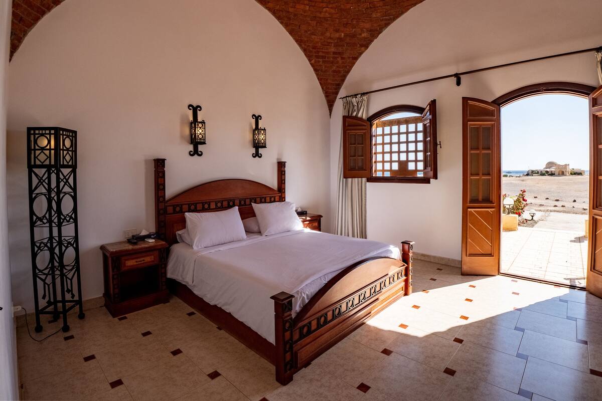 Een slaapkamer met een houten bed, twee wandlampen, een hoge vloerlamp en uitzicht op het strand door een open deur.