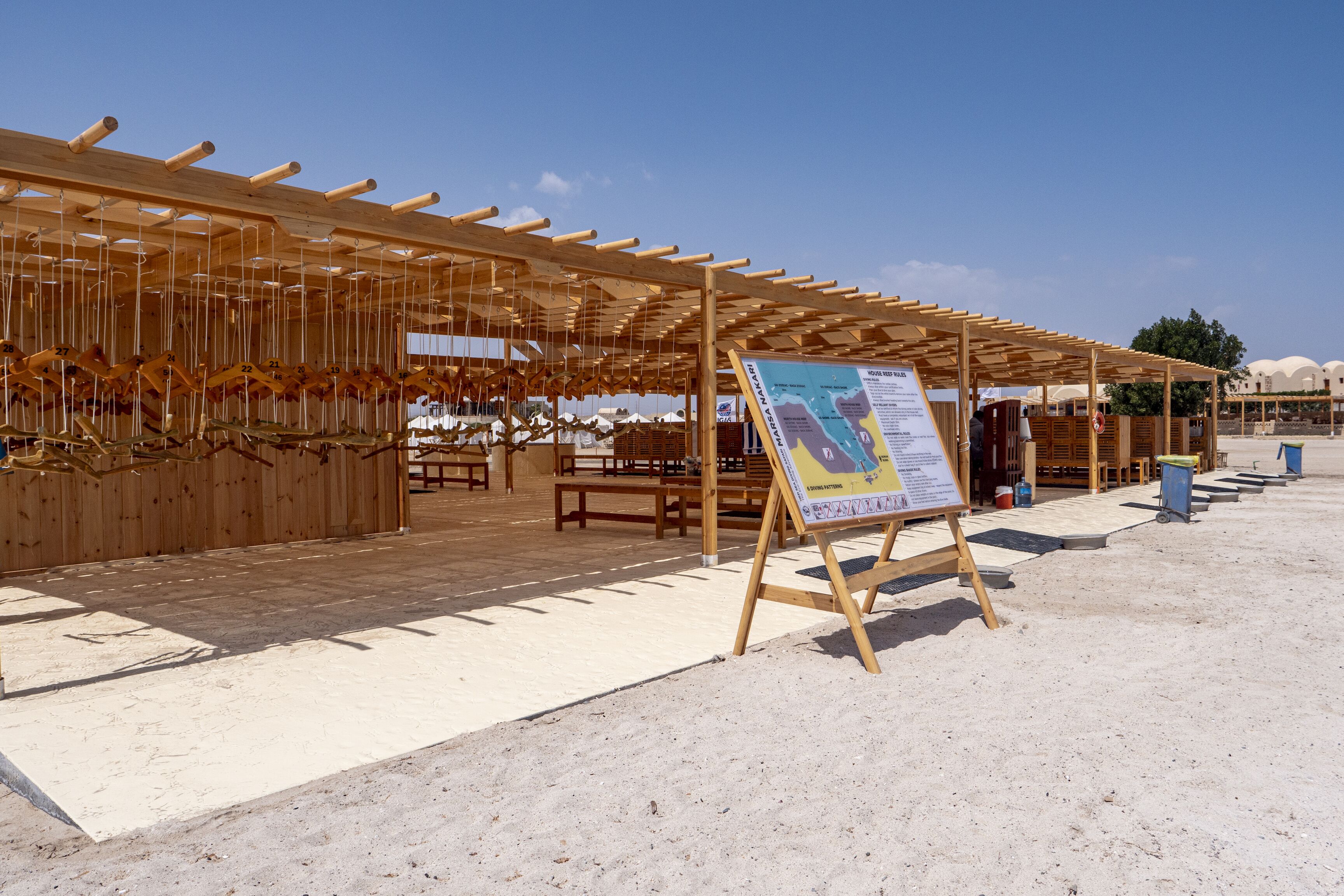 Une structure en bois formant une pergola, avec une enseigne, des bancs et un sol sablonneux.