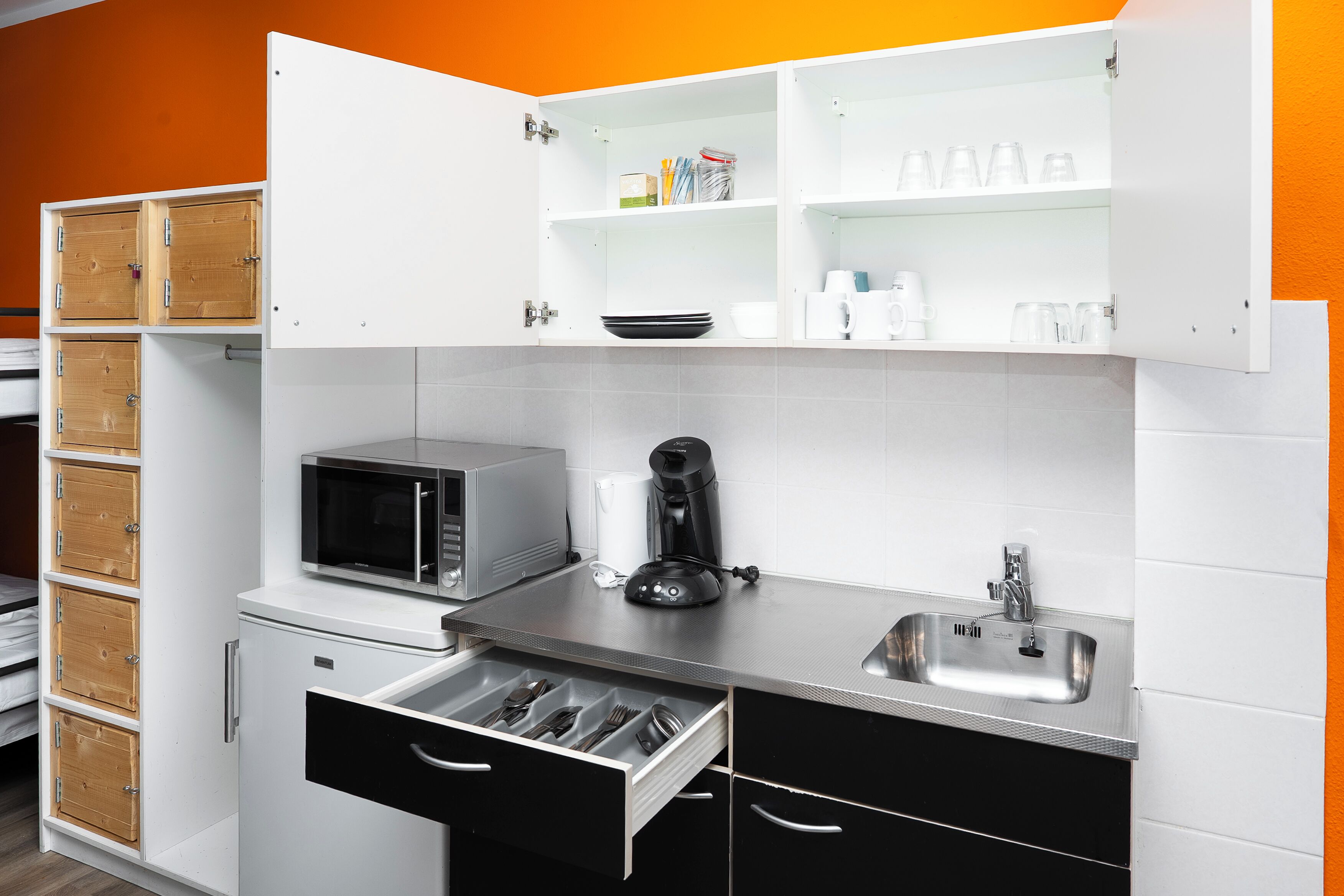 Una cocina compacta con armarios blancos, microondas, cafetera y lavamanos.