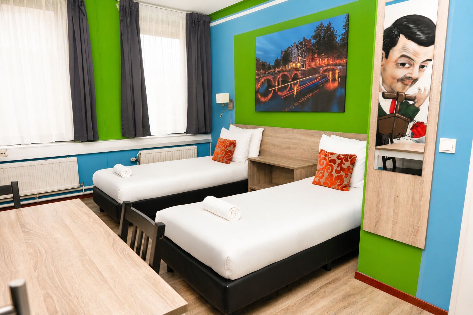 Ein Hotelzimmer mit zwei Betten, einem Holztisch, einem Stuhl, einem Fenster mit Vorhängen und einem Stadtbild.