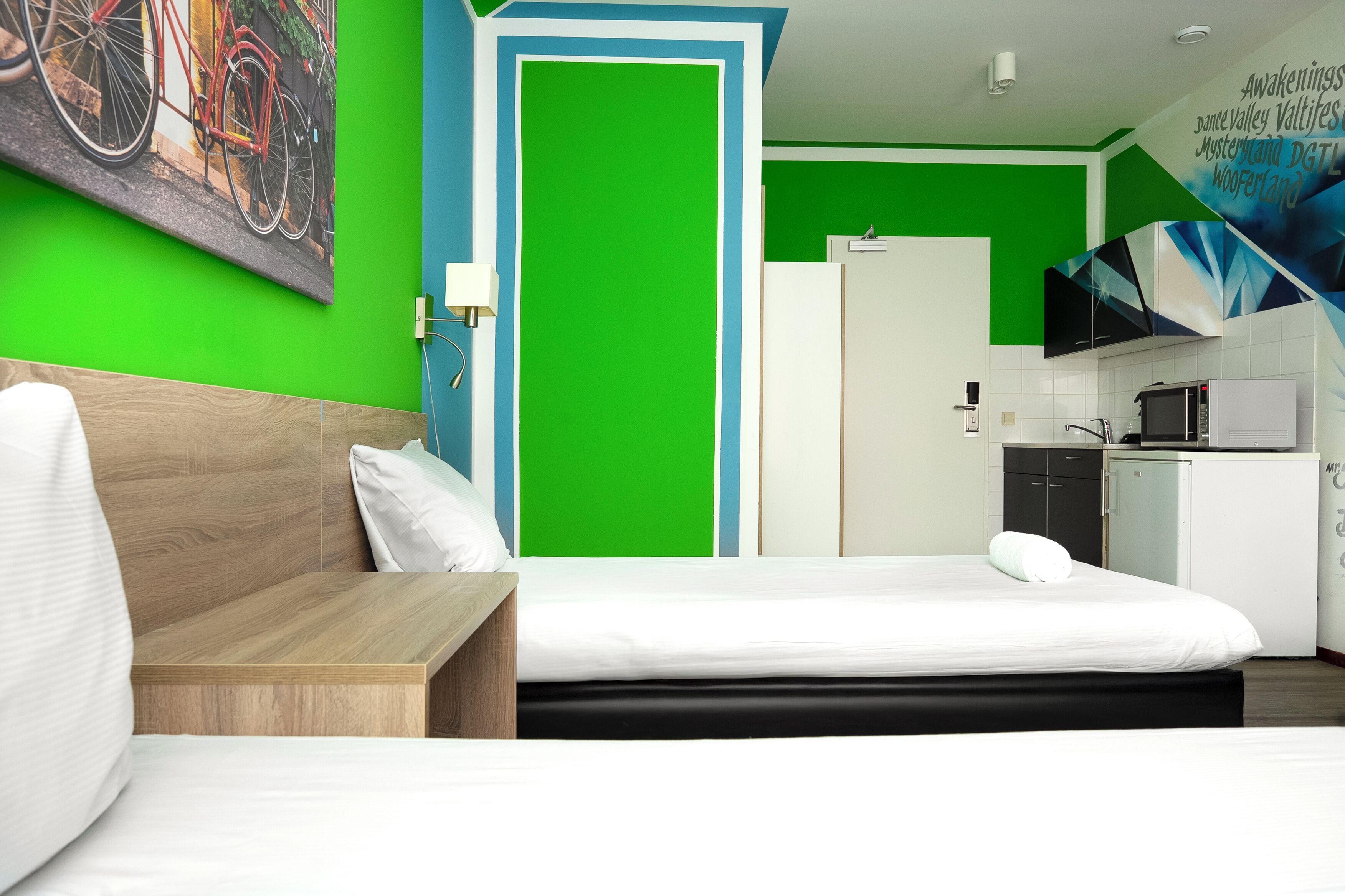 A hotel room with a bed, a wooden nightstand, a green wall, a white door, a kitchenette with a microwave, and a TV mounted on the wall.