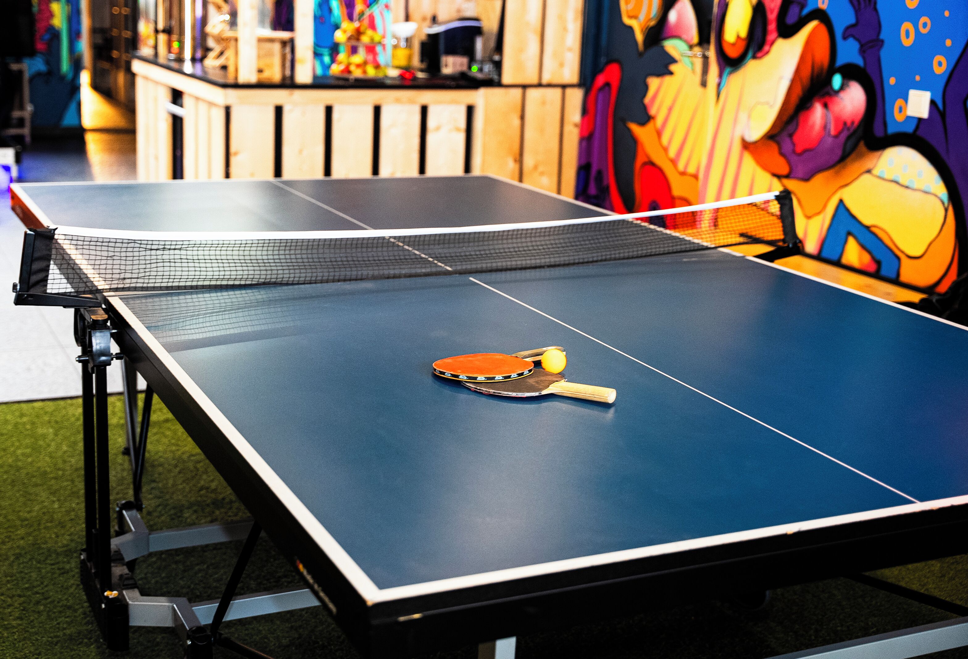 A ping pong table with a paddle and ball, set against a wall with colorful graffiti.