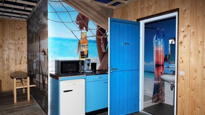 A compact room with a kitchenette, a blue door leading to a bathroom, and a mural of a beach scene. - Amsterdam Teleport Hotel (Amsterdam)