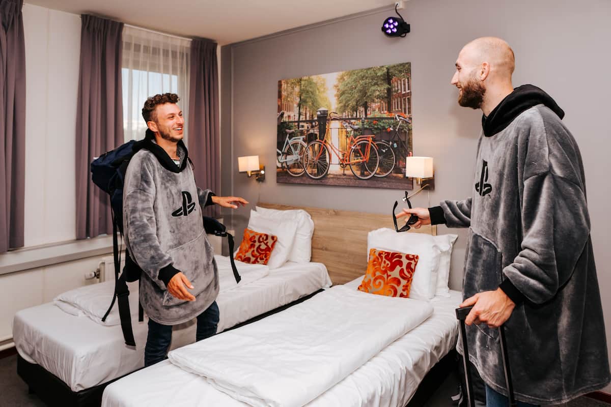 A hotel room with two beds, a painting of bicycles, and a lamp on the wall.