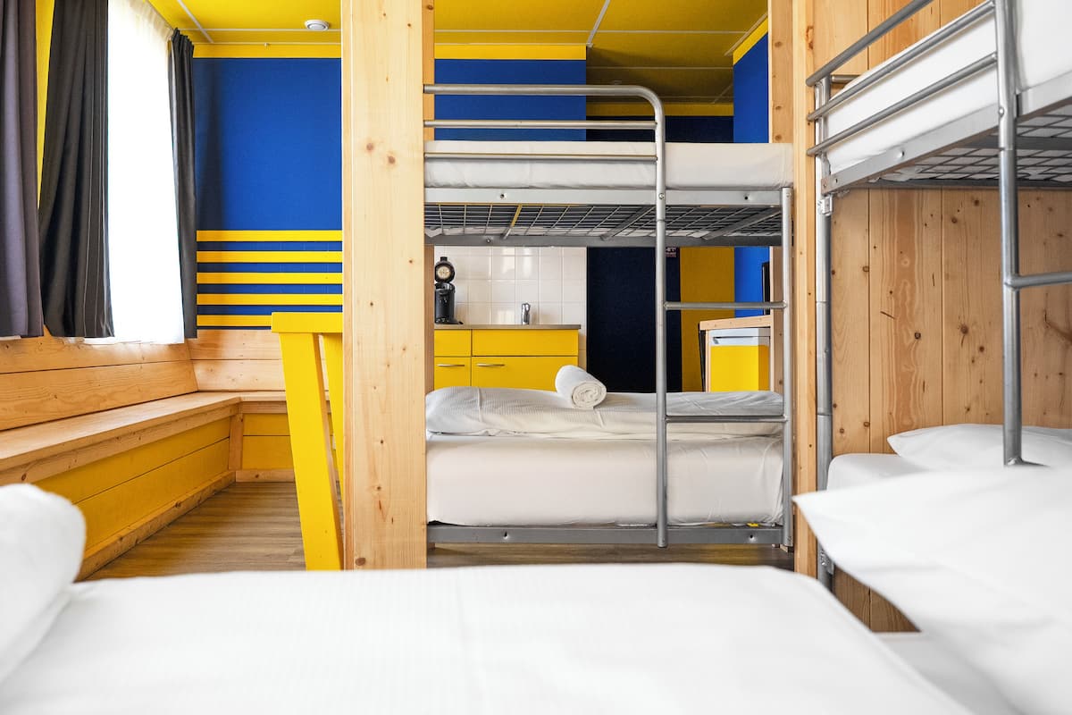 A bunk room with wooden walls, a kitchen area, and a window with curtains.