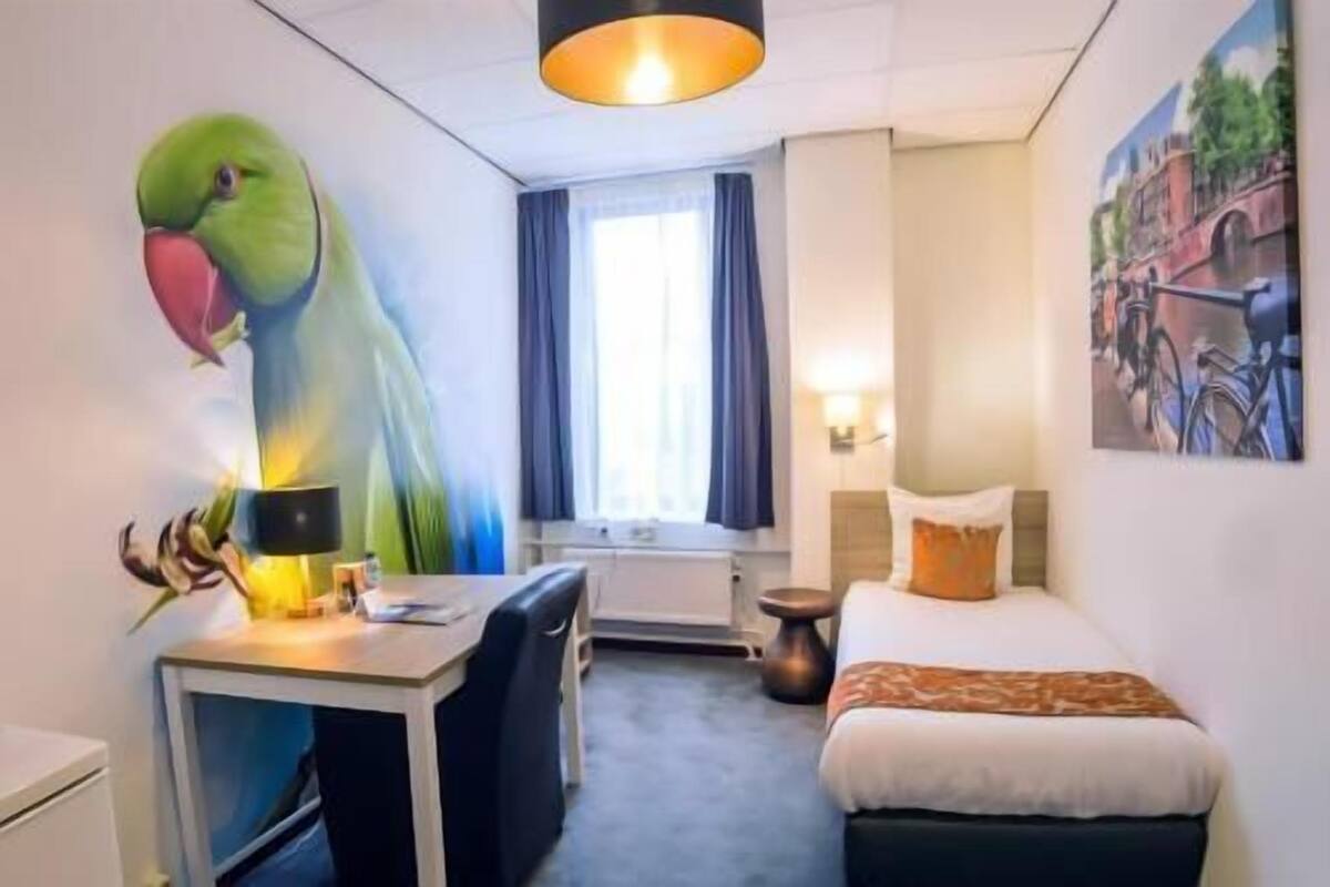 A hotel room with a bed, a desk with a chair, a lamp, a painting of a parrot, a window with curtains, and a wall-mounted light fixture.