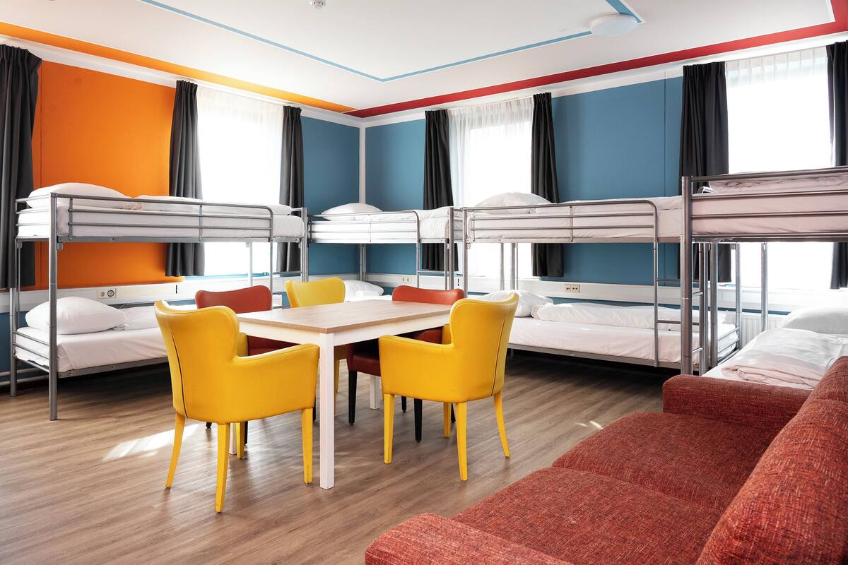 A dormitory room with bunk beds, a dining table with yellow chairs, and a red sofa.