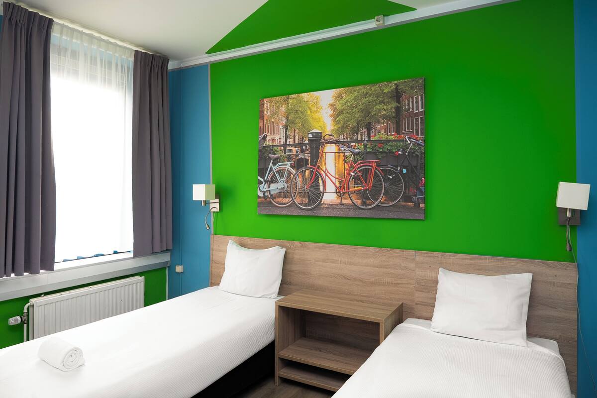 A hotel room with two beds, a green wall, a painting of bicycles, and a window with curtains.