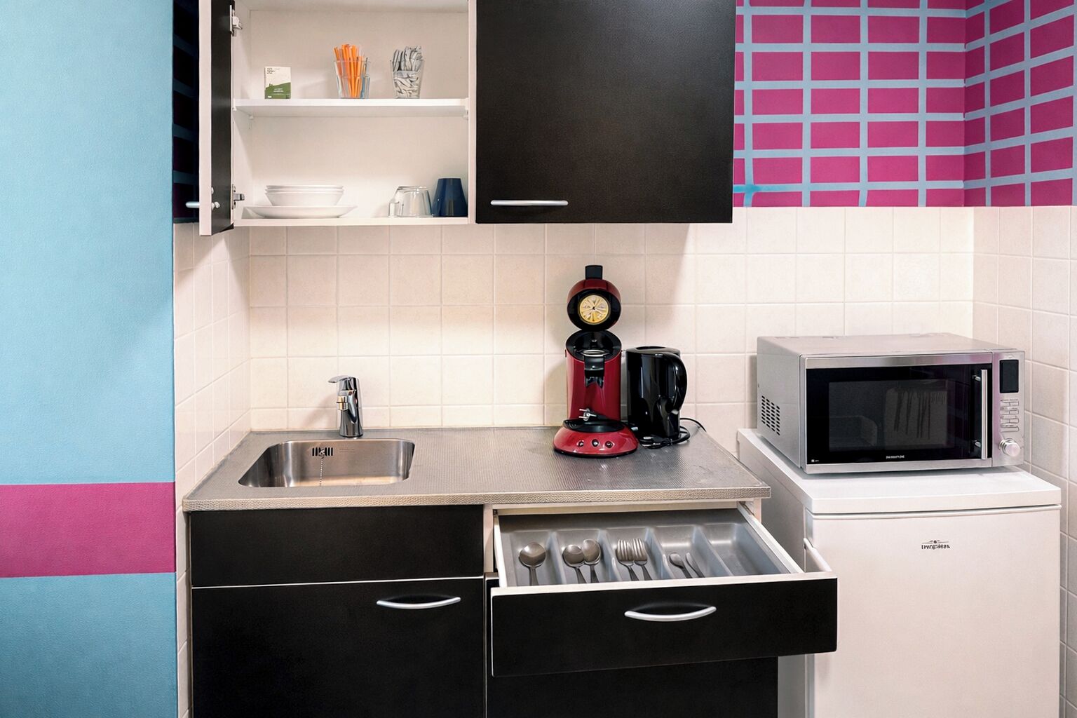 Una cucina compatta con lavandino, macchina da caffè, forno a microonde e un cassetto con utensili.