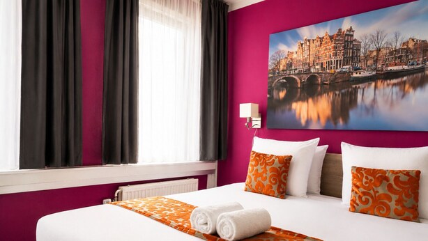 A hotel room with a large bed, a view of a cityscape, and a window with curtains.