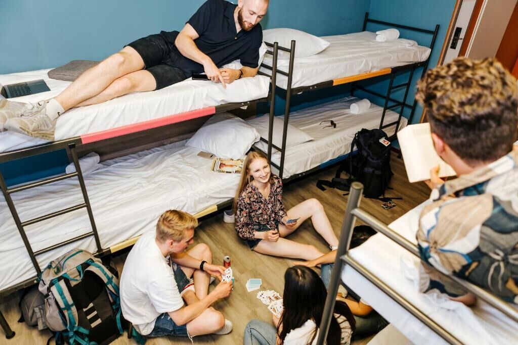 A dormitory room with bunk beds, a wooden floor, and a group of people sitting and interacting.