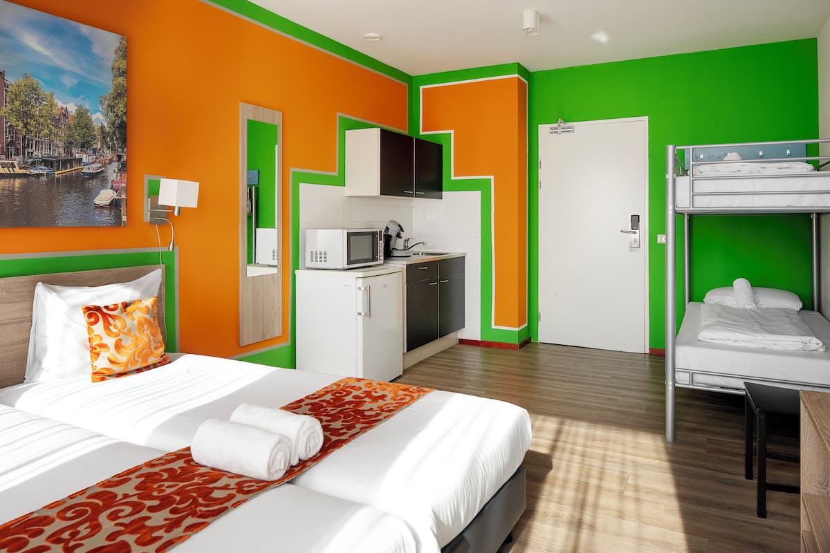 A hotel room with a bed, a bunk bed, a kitchenette, and a painting on the wall.