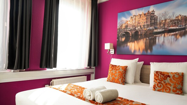 A hotel room with a bed, pillows, a painting of a cityscape, and a window with curtains. - Amsterdam Teleport Hotel (Amsterdam)