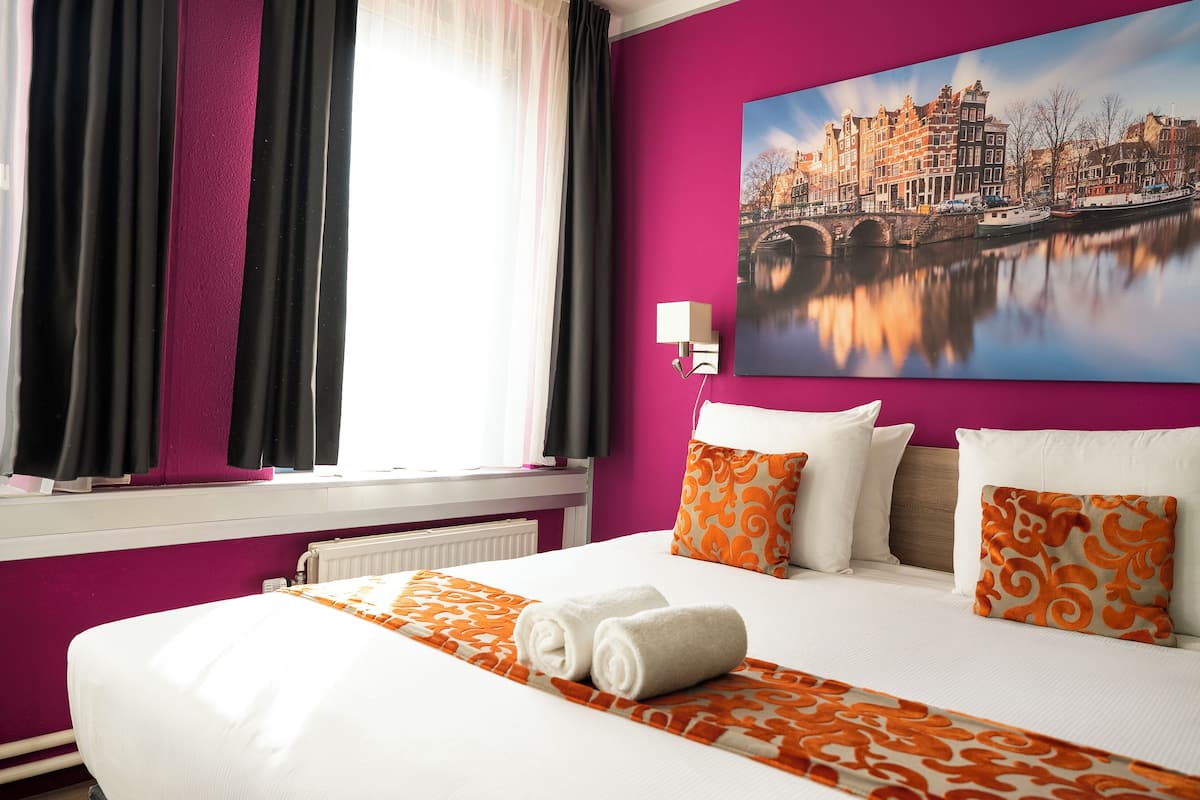 A hotel room with a bed, pillows, a painting of a cityscape, and a window with curtains.