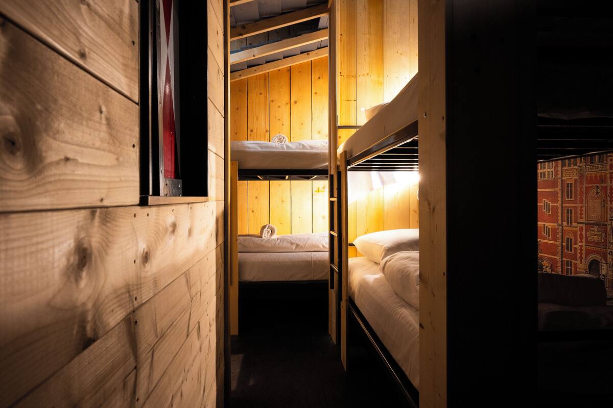 A narrow wooden room with bunk beds, a window with blinds, and a view of a building through the window.