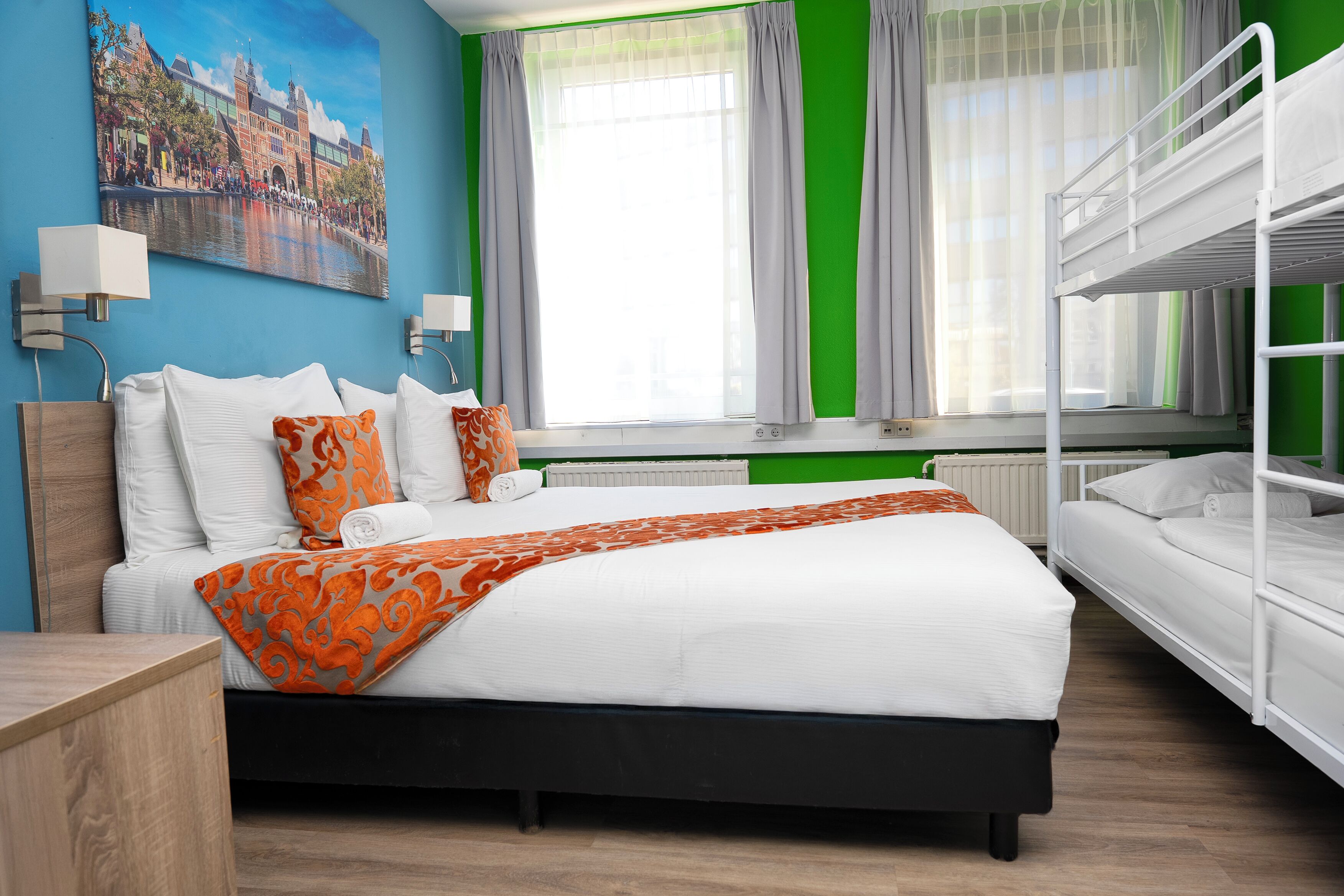 Una habitación de hotel con una cama grande, una cama litera, un cuadro de un paisaje urbano y una ventana con cortinas.