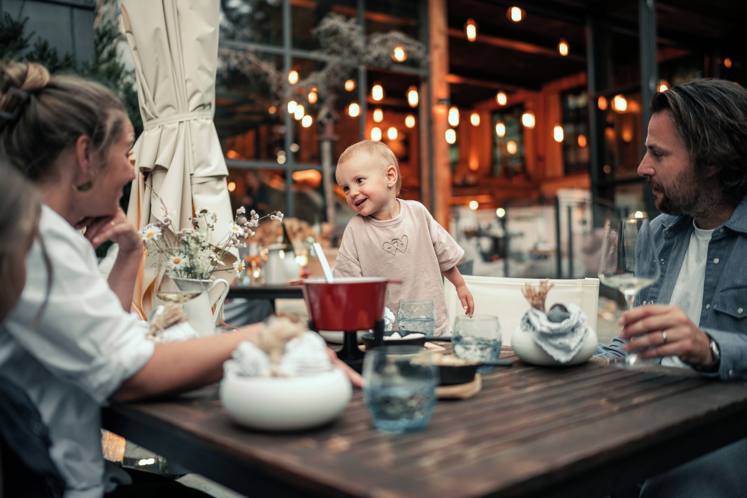 A family dining at a restaurant with a wooden table, various dishes, and a festive atmosphere with string lights.