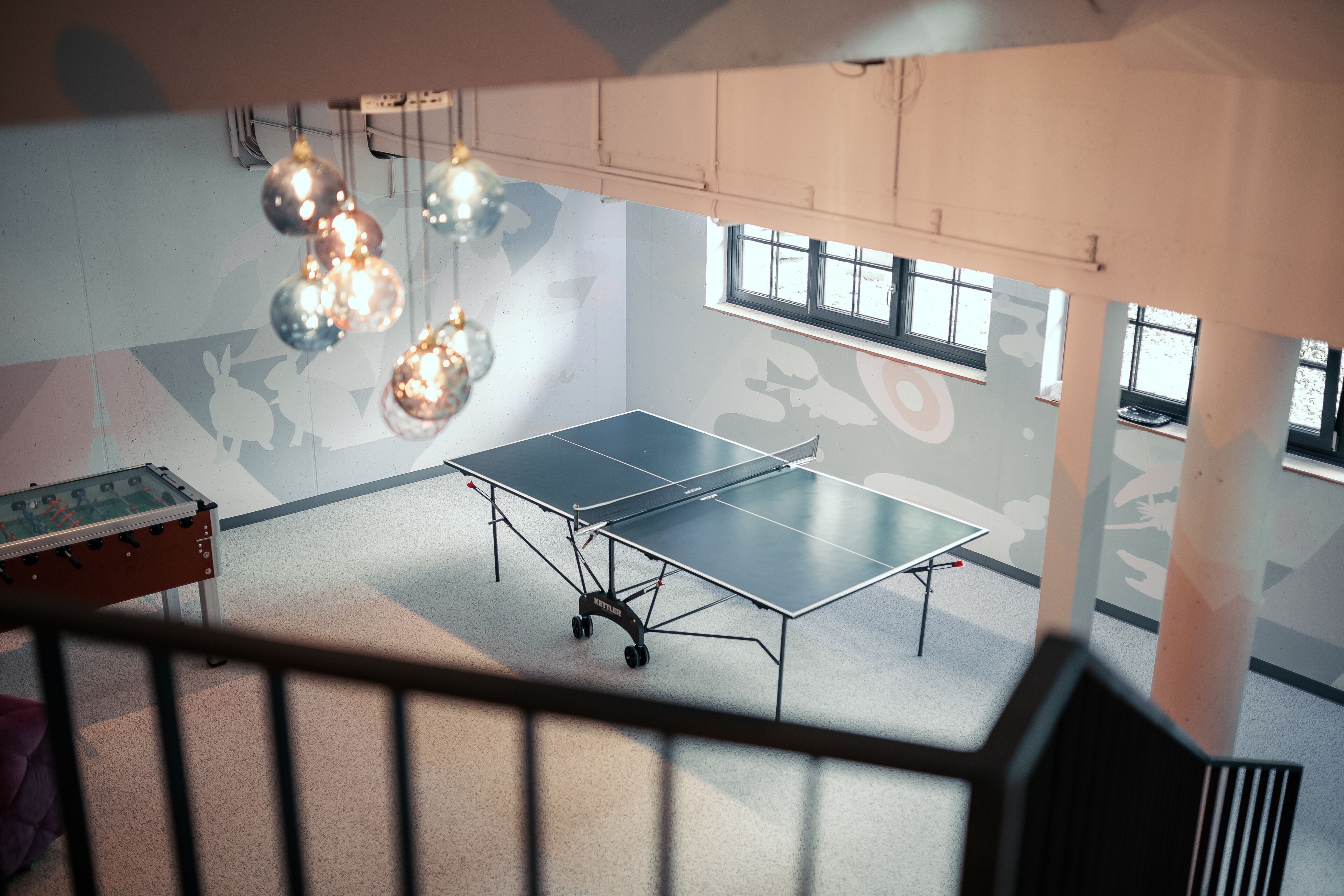 Una mesa de ping pong en una habitación con techo alto, ventanas amplias y arte abstracto en la pared.