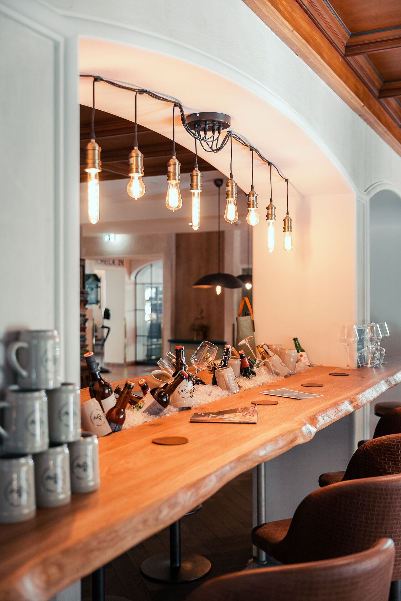 A bar area with a wooden counter, hanging light bulbs, and various bottles and glasses.