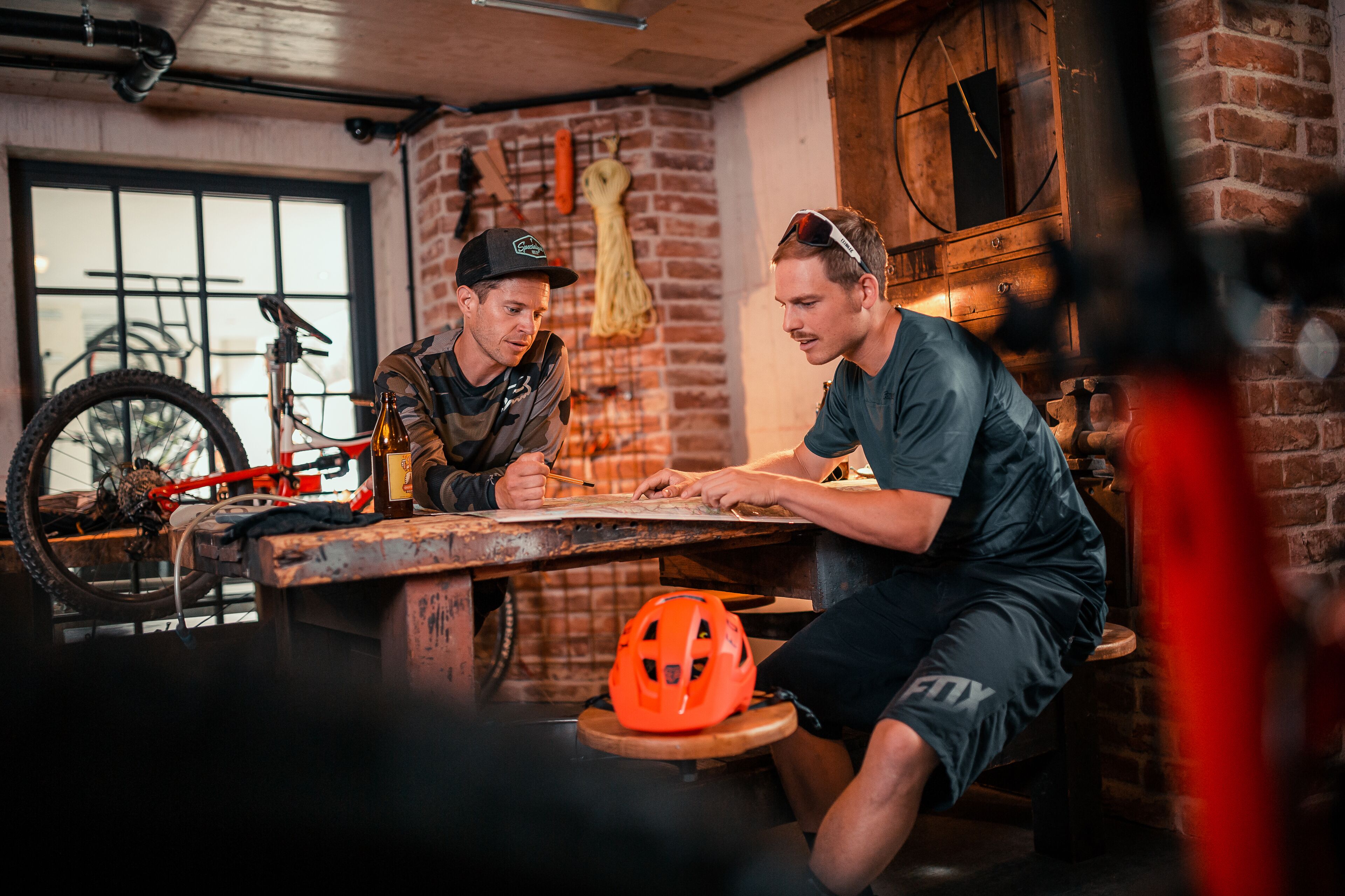 Zwei Männer sitzen an einem Holztisch in einem Raum mit Backsteinwänden, einem Fahrrad und einem Helm auf dem Boden.