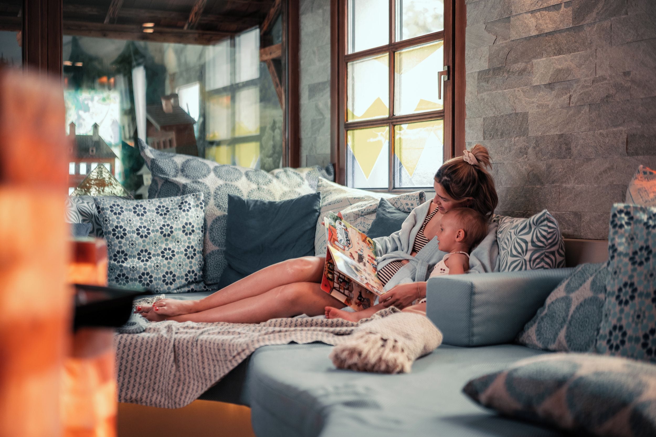 A person reading a book on a sofa with patterned cushions and a window in the background.