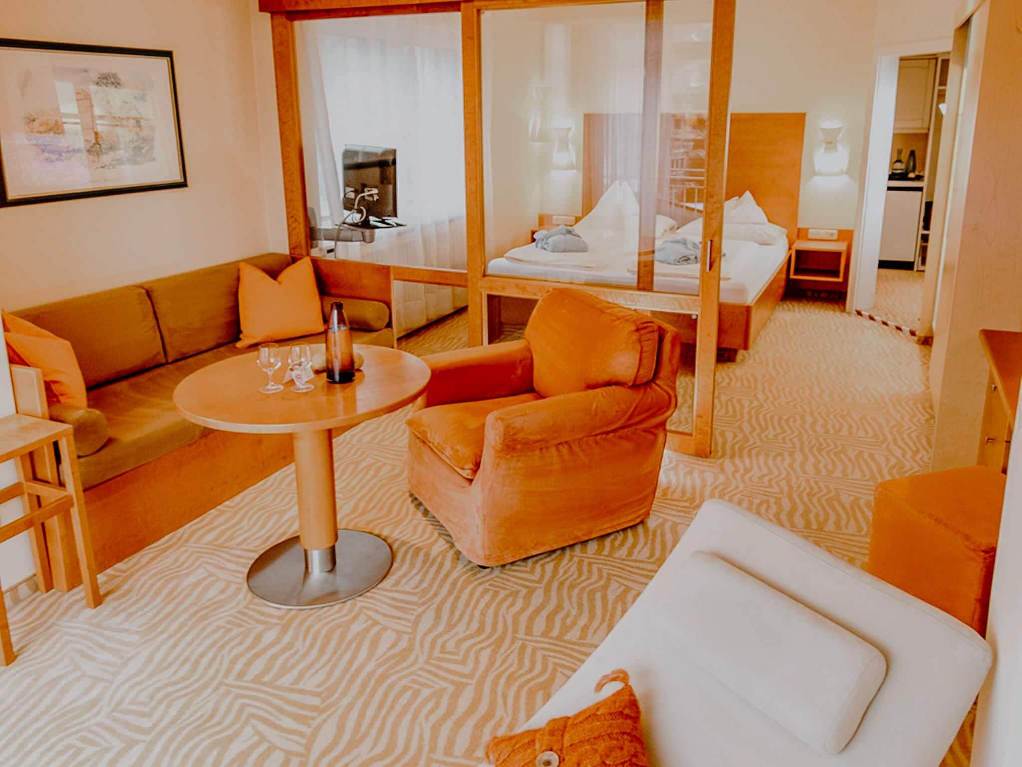 Habitación de hotel con sofá, sillón y una pequeña mesa con vasos. Se observa una cama con dosel y una mesita de noche con lámpara.