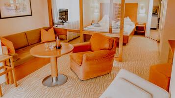 Habitación de hotel con sofá, sillón y una pequeña mesa con vasos. Se observa una cama con dosel y una mesita de noche con lámpara.