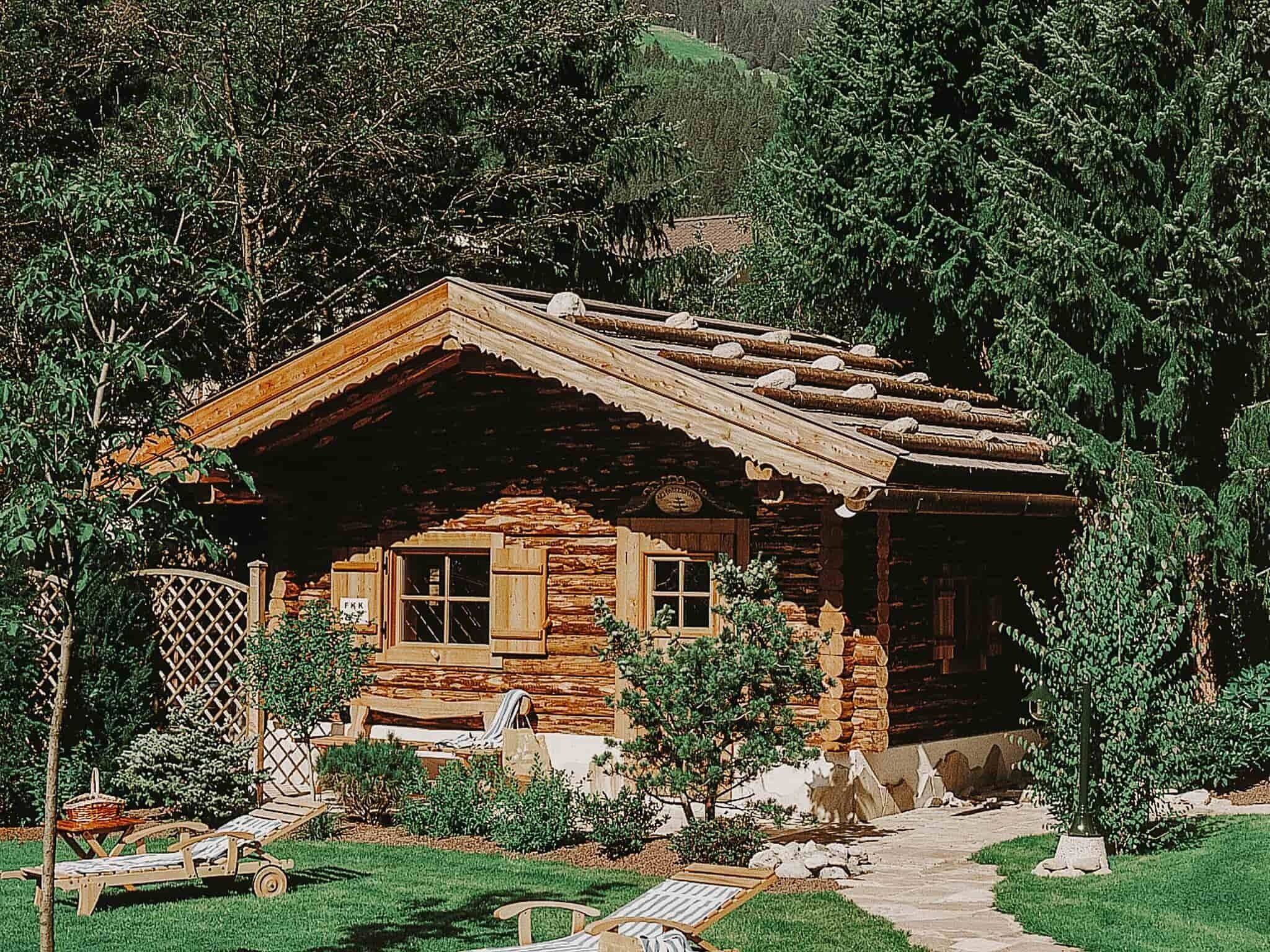 A rustic log cabin with a porch, surrounded by greenery and a clear path leading to the entrance.