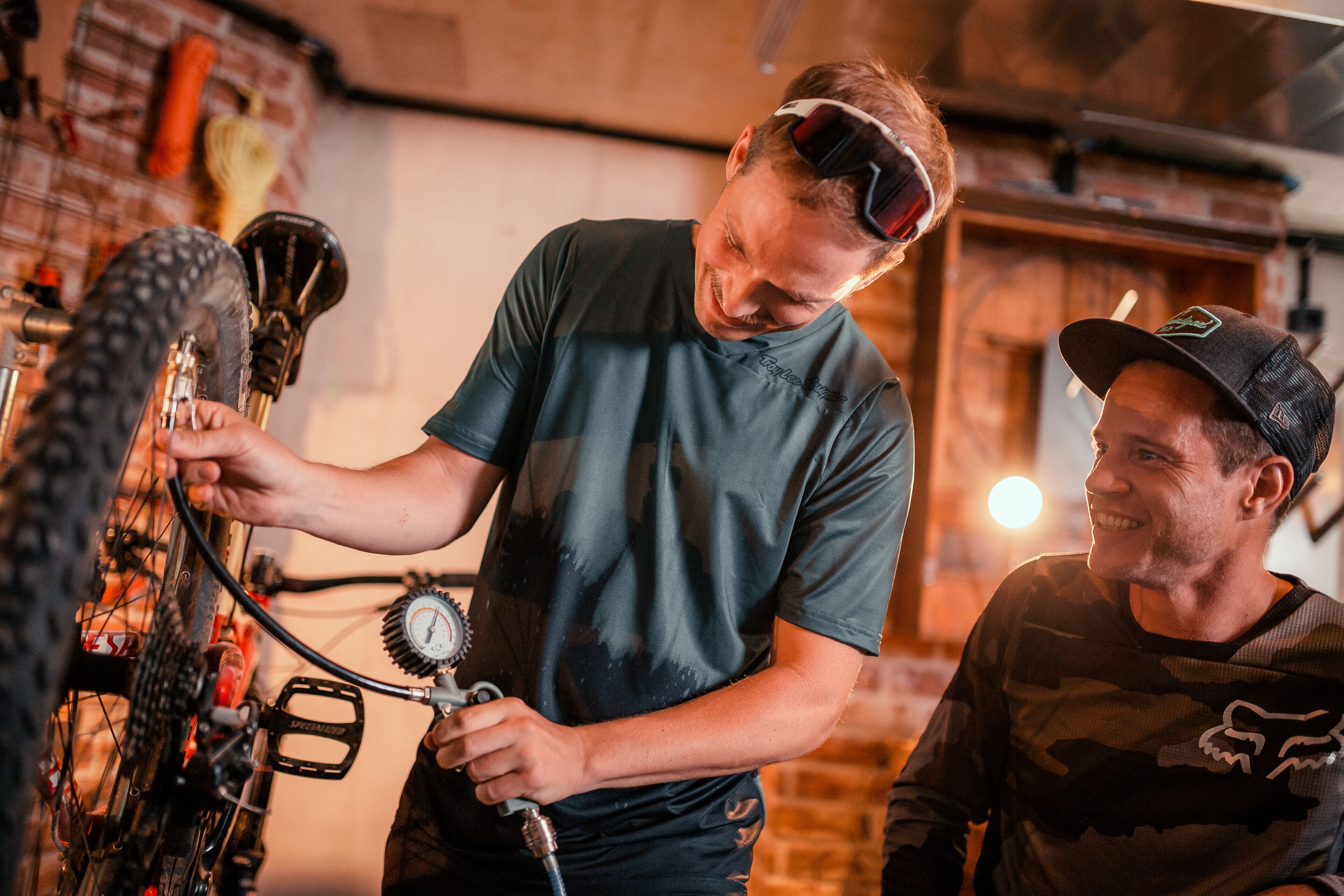 Two people working on a bicycle in a workshop with brick walls and tools.
