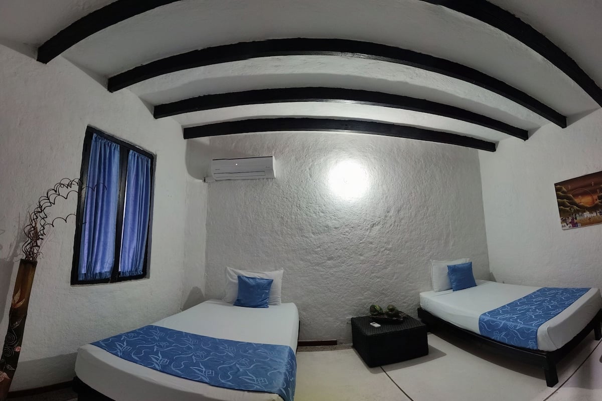 Ein Zimmer mit zwei Betten, einem Fenster mit blauen Vorhängen, einer Klimaanlage und einem kleinen schwarzen Tisch.