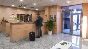 Ein Hotel-Empfangsbereich mit Empfangstresen, einem Blumentopf, einem Tisch mit Broschüren und einer Glastür zum Außenbereich.