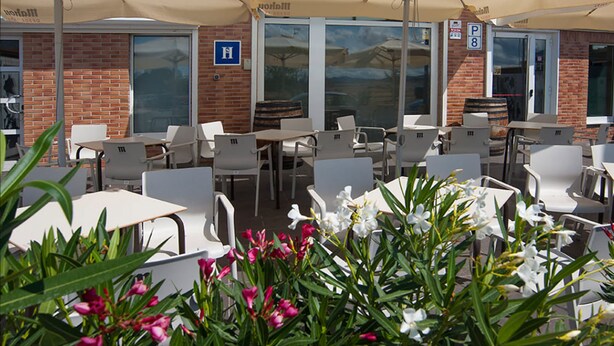 Ein Straßencafé mit weißen Tischen und Stühlen, beigen Sonnenschirmen und einer Ziegelwand.