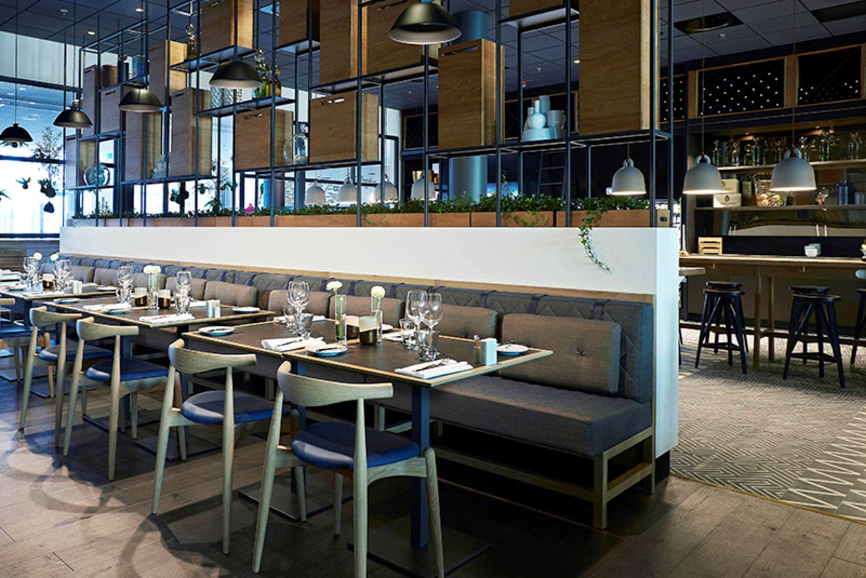 A modern restaurant with multiple dining tables and booths, a bar area, and a high ceiling with pendant lighting.