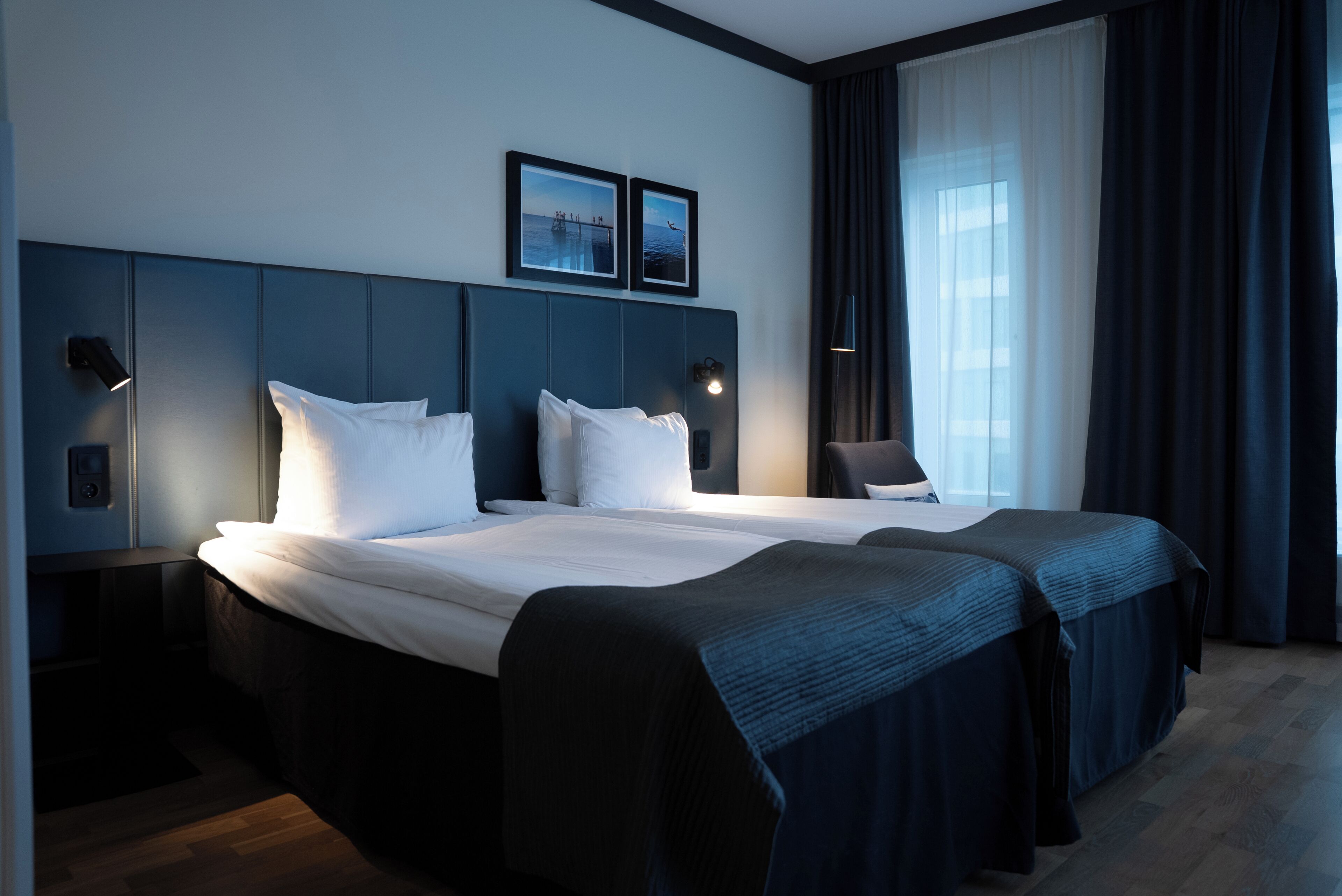 A hotel room with a large bed, two bedside tables, a headboard with wall art, and a window with curtains.