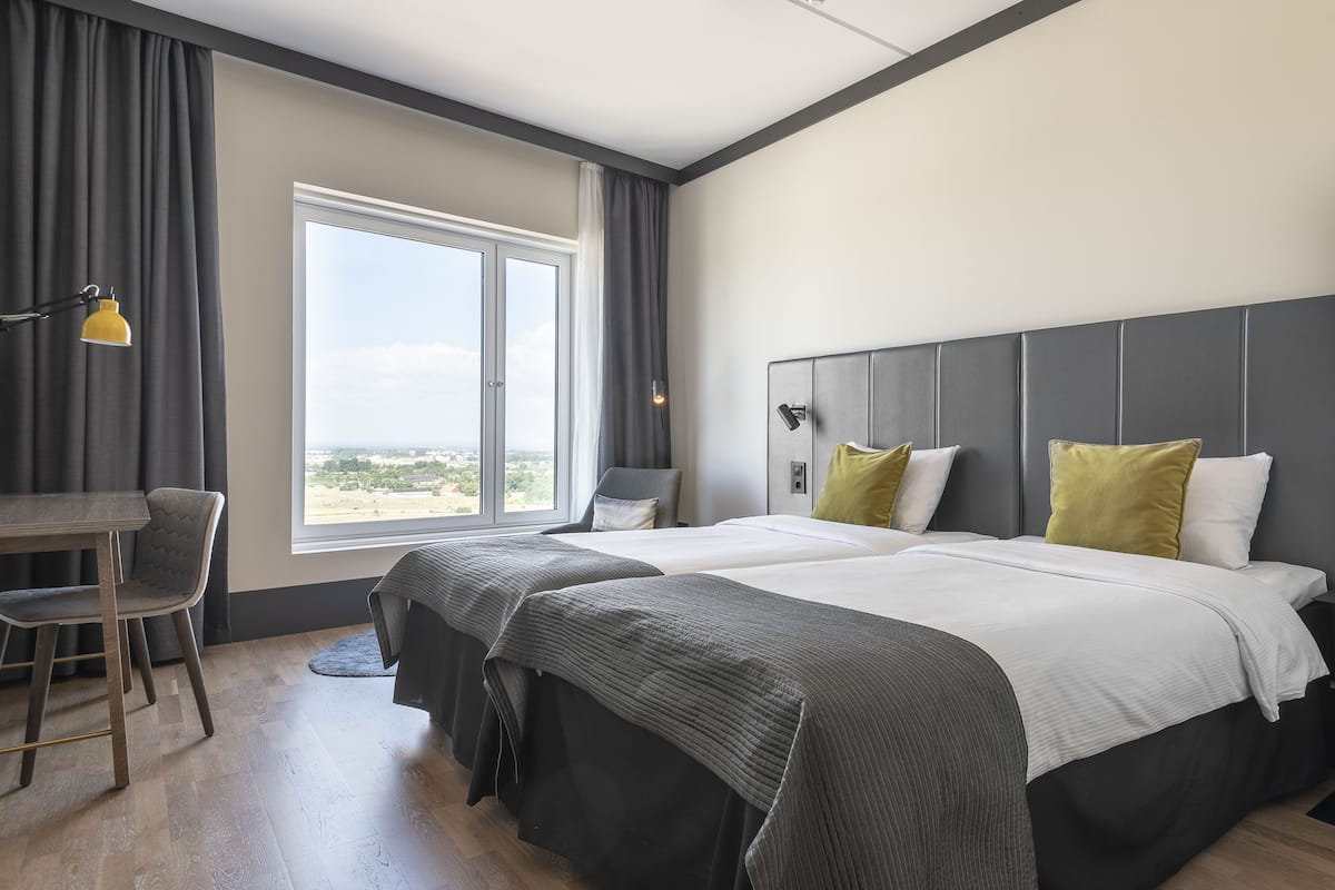 A hotel room with two beds, a desk, and a large window.