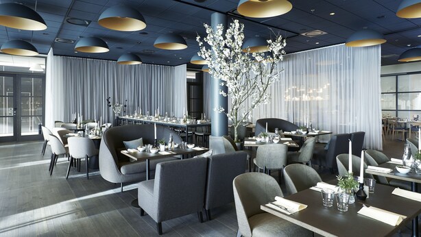 Un restaurant moderne avec un plancher en bois foncé, des chaises à l’assise rembourrée de couleur grise et des tables recouvertes de nappes blanches et décorées de bougies.