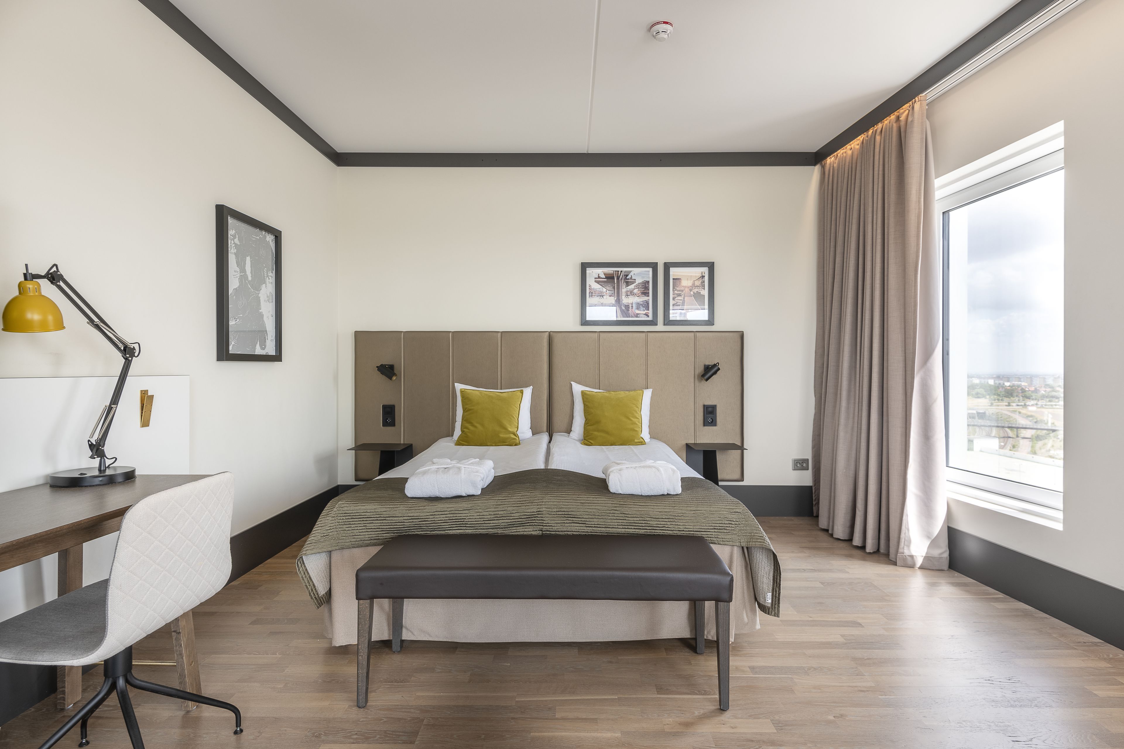 A modern hotel room with a large bed, a desk, a chair, and a view of the city.