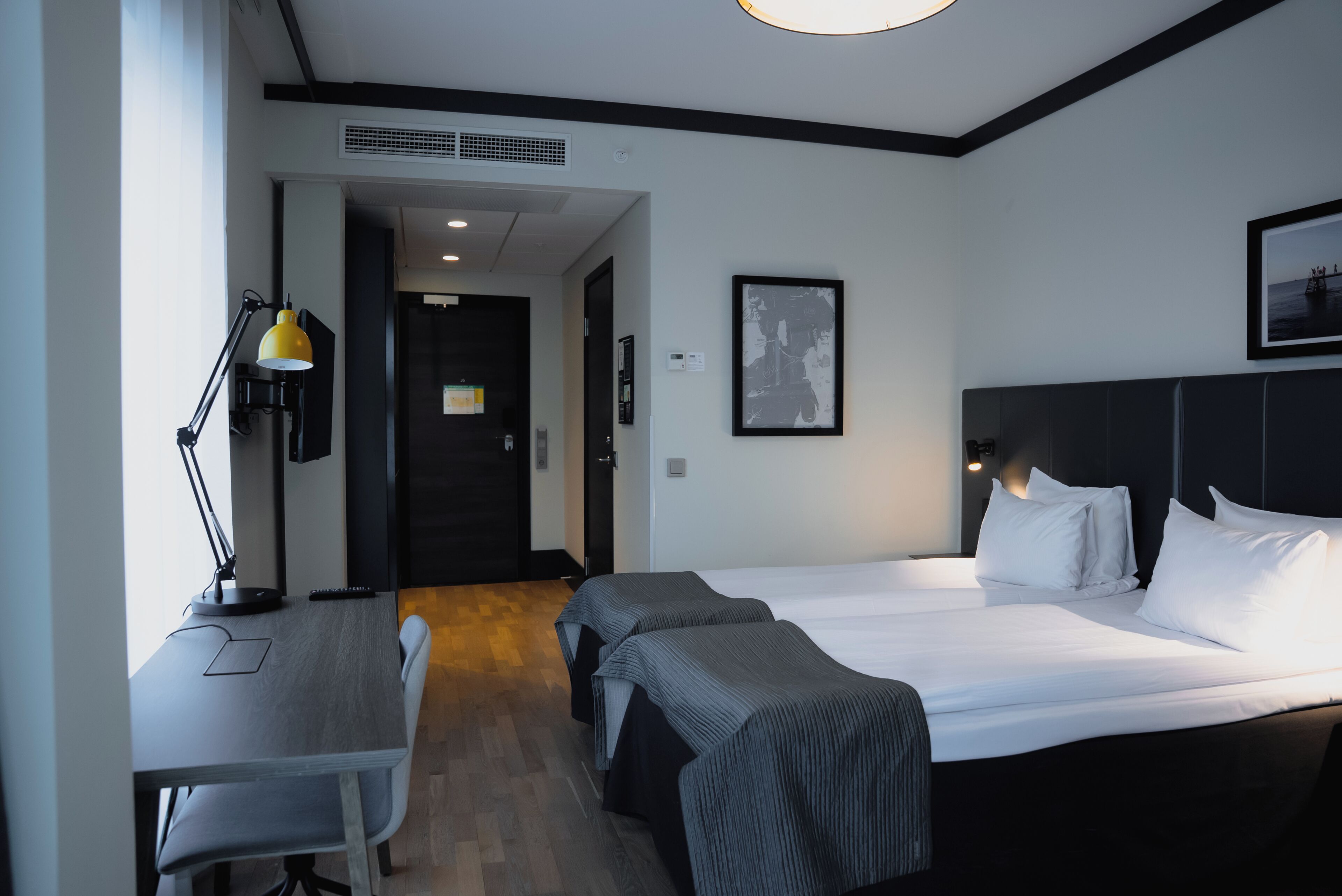 Ett hotellrum med en säng, sängbord, ett skrivbord, en lampa och en inramad tavla på väggen.