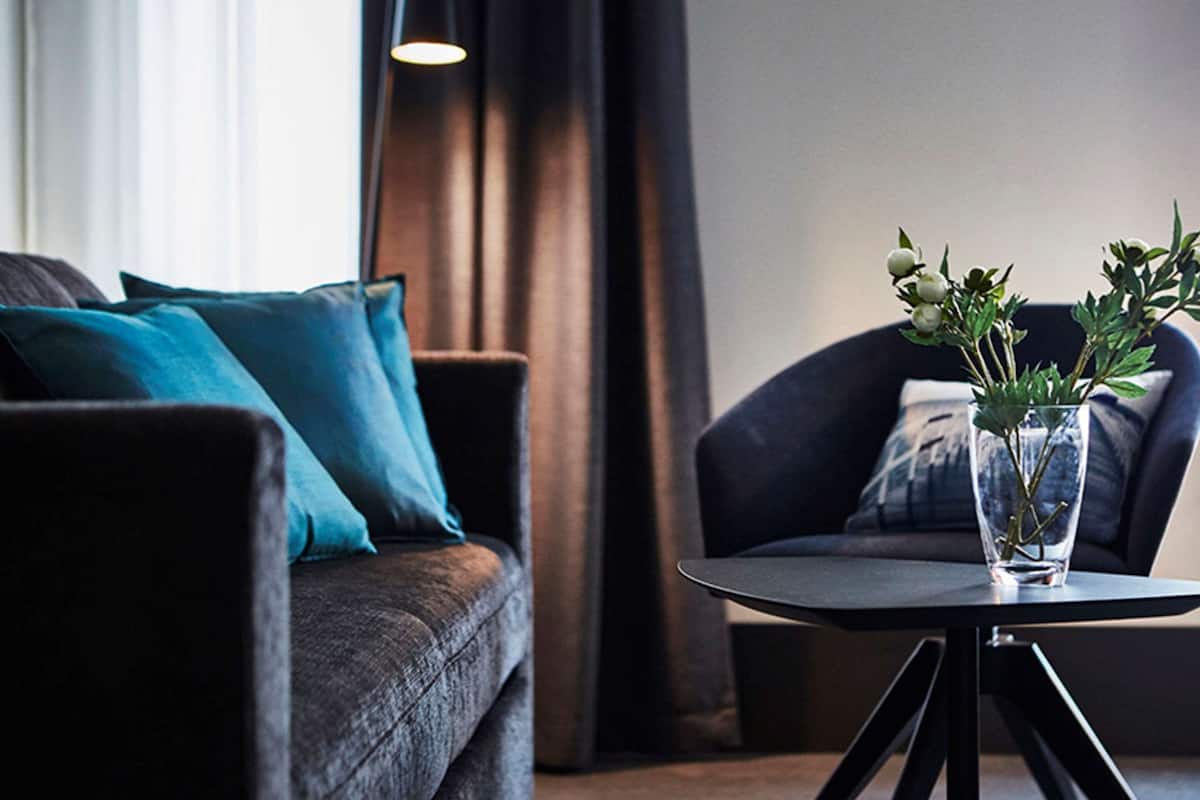 A modern living room with a dark sofa, teal cushions, a dark armchair, and a small round table with a clear vase containing green plants.