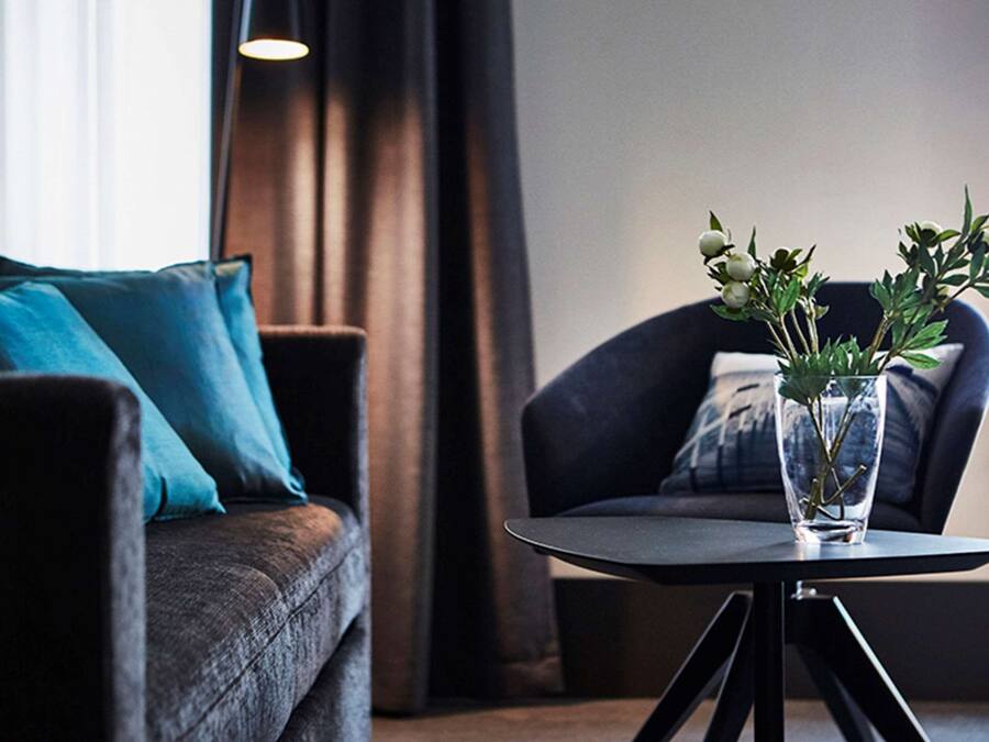 Una sala moderna con un sofá oscuro, cojines azul verdosos, un sillón oscuro y una pequeña mesa redonda con un florero transparente que contiene plantas verdes.