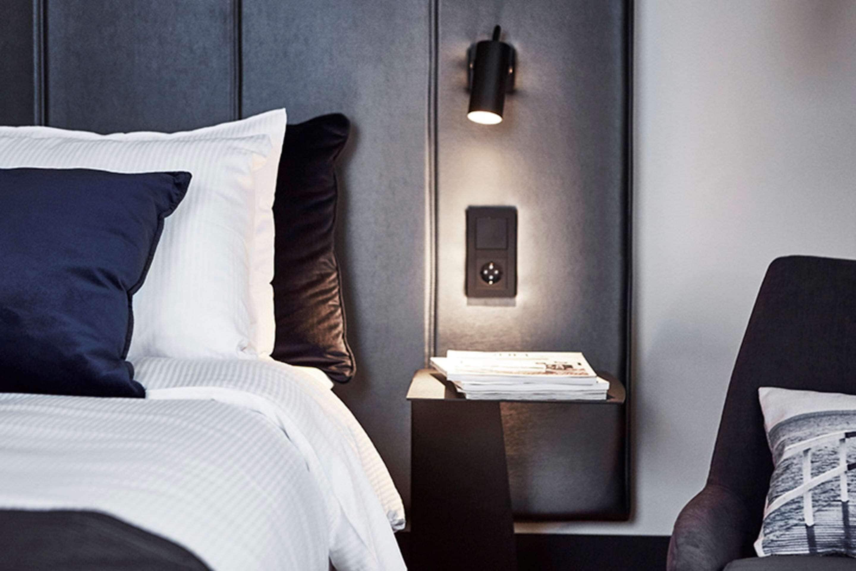 A neatly made bed with white and navy pillows, a bedside table with a lamp, and a chair with a cushion.