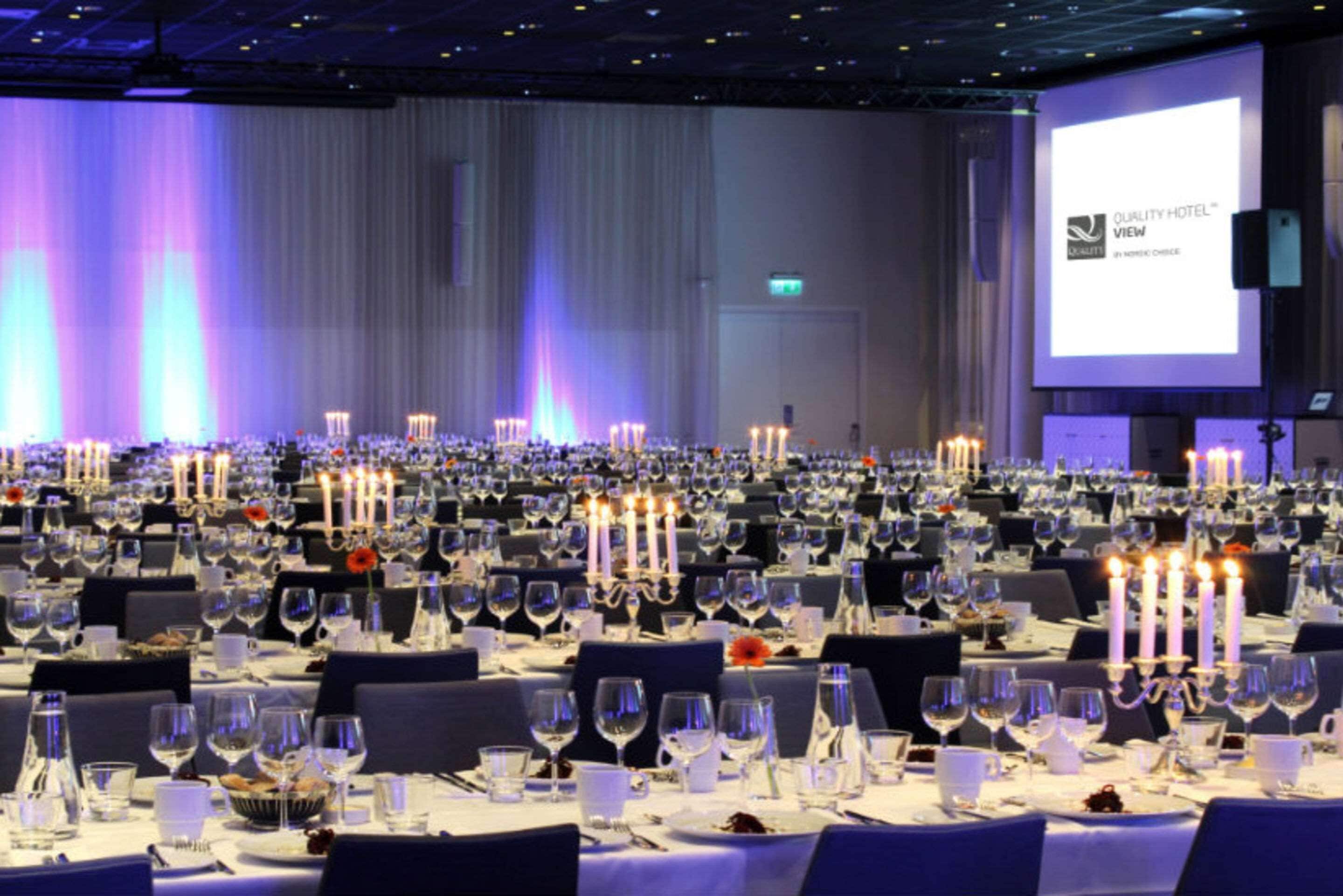 Un salón de banquetes con mesas dispuestas para un evento formal, con una pantalla grande mostrando el nombre del hotel.