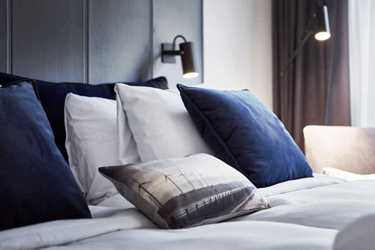 A neatly made bed with multiple pillows, a wall-mounted light fixture, and a window with curtains.