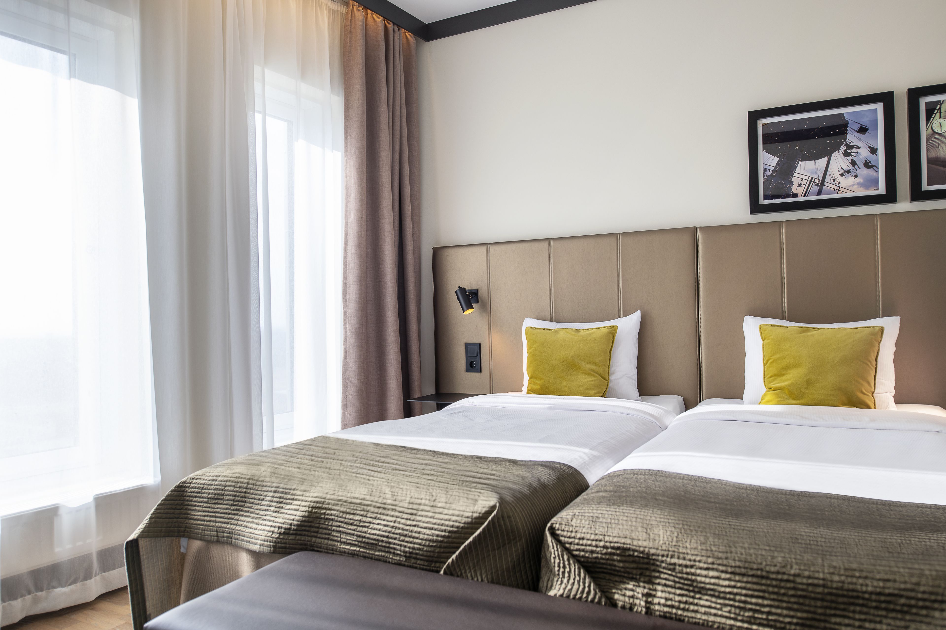 A hotel room with two beds, a large window with sheer curtains, and a wall-mounted lamp.