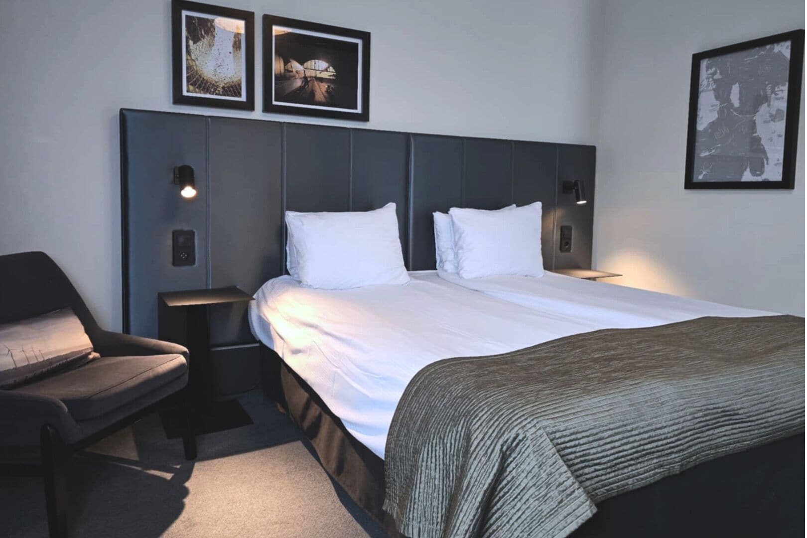 A hotel room with a bed, a chair, a nightstand, and framed pictures on the wall.