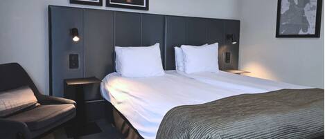 A hotel room with a bed, a chair, a nightstand, and framed pictures on the wall.